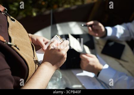 Kaukasische junge Erwachsene Frau mit Notizblock, die Bestellung von einem kaukasischen Mann mittleren Alters nimmt, der am Tisch sitzt, mit Laptop und Stift hält, im Freien Café-Einstellung, Hände sichtbar Stockfoto