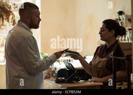 Kaukasischer junger erwachsener Mann, der kontaktlos mit dem Smartphone bezahlt, während hispanische junge Erwachsene Frau, die Kartenleser am Schalter im Café hält, beide konzentrierten sich auf Transaktionen Stockfoto