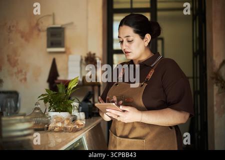 Junge erwachsene hispanische Frau, die hinter der Theke steht und ein digitales Tablet verwendet, sich auf den Bildschirm konzentriert, während sie Bestellungen im Café oder in der Bäckerei verwaltet, Schürze trägt, Haare zurückgebunden Stockfoto
