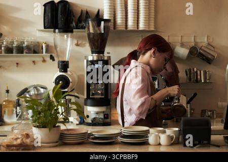 Junge Erwachsene weiße Frau mit roten Haaren bereitet Kaffee hinter der Theke in einem modernen Café vor, wobei professionelle Kaffeegeräte verwendet werden und sich darauf konzentriert, Getränke in eine Tasse zu gießen Stockfoto