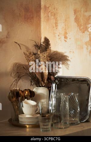Stillleben-Arrangement mit getrocknetem Pampasgras und Laub in Keramikvase neben stapelbaren Tellern, Tassen, Besteck, Gläsern und Serviertablett aus Metall auf Holzoberfläche Stockfoto