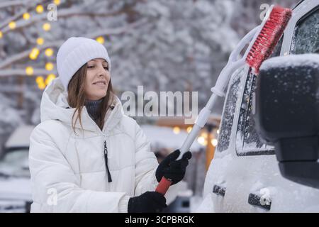 Eine 30- bis 35-jährige Frau in weißer Jacke und Hut mit einem Lächeln im Gesicht reinigt den festsitzenden Schnee und das Eis von ihrem Auto und macht sich bereit Stockfoto