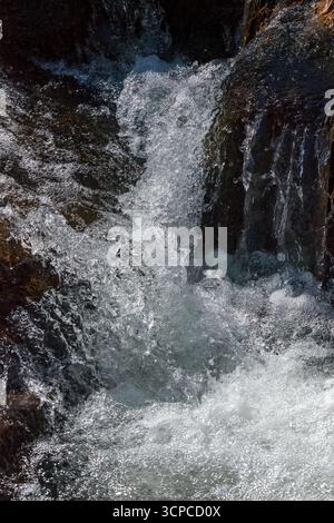 Nahaufnahme eines schnell fließenden Baches, der über dunkle Felsen stürzt. Das Sonnenlicht reflektiert das Wasser und erzeugt glänzende Highlights und dynamische Texturen in einem Stockfoto