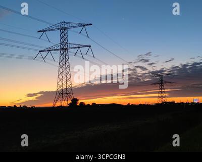 Elektrische Pylonen, die bei Sonnenaufgang über einem offenen Feld stehen Stockfoto