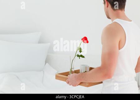 Mann, der im hellen Schlafzimmer steht und Holztablett mit Rose und Becher neben dem Bett hält, Kopierraum Stockfoto