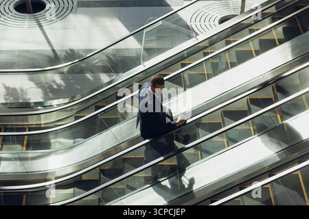 Geschäftsmann packt Lederakkoffer, steigt auf Stahltreppen im Glasatrium, wirft Schatten Stockfoto