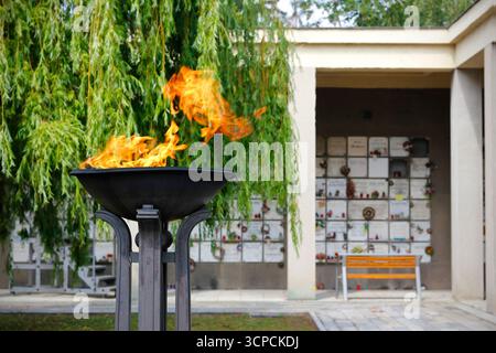 Ewiges Feuer brennt in schwarzer Metallschale im Krematoriumhof mit Urnenwand Kolumbarium und Gedenkbank im Hintergrund, Symbol der Erinnerung. Stockfoto