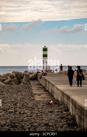 Eine Gruppe von Menschen wird auf einem Betonpier neben einem felsigen Wellenbrecher mit einem markanten grün-weiß gestreiften Leuchtturm V beobachtet Stockfoto