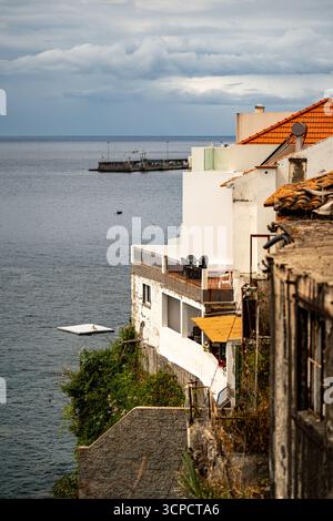 Eine Person entspannt sich auf einer weißen schwimmenden Plattform, während eine andere im Meer schwimmt, mit Blick auf die Klippen von Gebäuden mit Balkonen und Terrakottadächern. Stockfoto