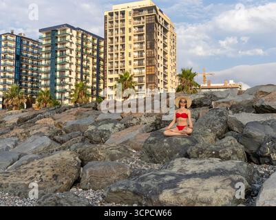 Attraktive Frau mit langen Haaren, rotem Bikini und Strohhut, am Strand, sitzend auf einem großen Felsen, meditierend. Stockfoto