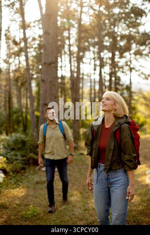 Zwei Wanderer erkunden einen ruhigen Wald mit hohen Bäumen und warmer Sonne. Die Frau sieht glücklich und entspannt aus, während der Mann dicht hinter sich her folgt. Stockfoto