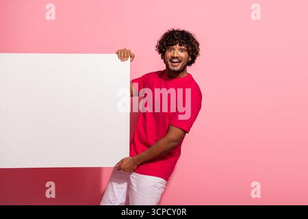 Glücklicher junger Mann, der ein weißes Schild vor rosa Hintergrund hält, lässige Kleidung trägt und Aufregung ausstrahlt Stockfoto