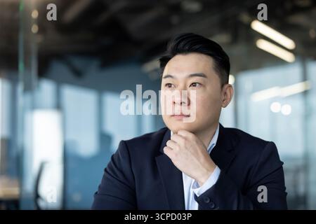 Nahaufnahme eines jungen seriösen asiatischen Mannes in einem Business-Anzug, der im Büro ist und nachdenklich aus dem Fenster schaut. Stockfoto