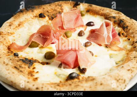 Blick von oben auf eine Gourmet-Pizza mit geschmolzenem Mozzarella, Schinkenscheiben und einer Mischung aus grünen und schwarzen Oliven. Stockfoto