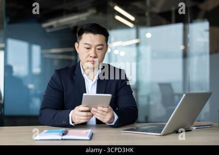 Asiatischer junger männlicher Geschäftsmann, der im Büro an einem Tisch mit einem Laptop sitzt und konzentriert ein Tablet benutzt. Stockfoto