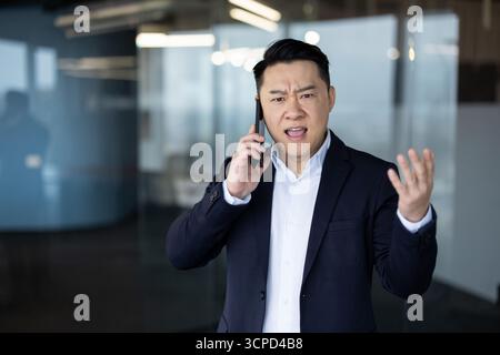 Porträt eines wütenden jungen asiatischen Geschäftsmannes, der im Büro steht und emotional telefoniert. Stockfoto