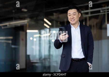Ein lächelnder asiatischer Geschäftsmann in einem Anzug schaut sich sein Handy in einer Büroumgebung an. Er scheint Nachrichten oder Neuigkeiten zu prüfen. Stockfoto