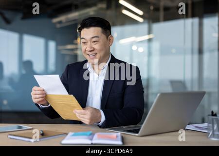 Glücklicher junger asiatischer Mann, der im Büro an seinem Schreibtisch sitzt und lächelt, während er einen erhaltenen Brief liest. Stockfoto