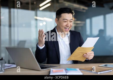 Glücklicher junger asiatischer Mann, der an seinem Schreibtisch im Büro sitzt und einen erhaltenen Brief liest. Stockfoto