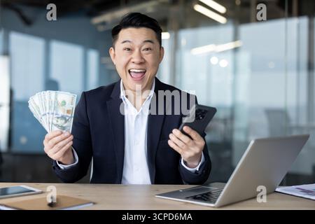 Porträt eines jungen asiatischen Mannes in einem Anzug, der im Büro an einem Schreibtisch sitzt, einen Geldschein und ein Telefon hält und mit einem Lächeln auf die Kamera blickt. Stockfoto