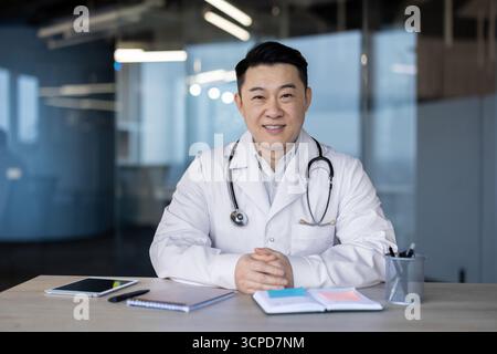 Porträt eines lächelnden jungen asiatischen Arztes in einem weißen Mantel und mit einem Stethoskop, das an seinem Schreibtisch sitzt und in die Kamera lächelt. Stockfoto