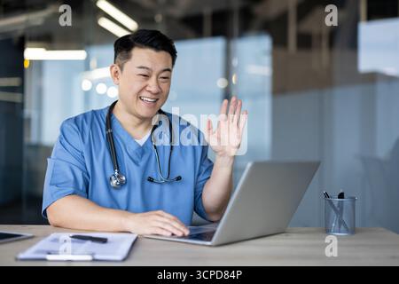 Asiatisch lächelnder junger männlicher Arzt, Student sitzt im Krankenhaus am Schreibtisch in blauer Uniform, grüßt und spricht auf Laptop-Kamera. Stockfoto