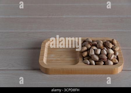 Blick von oben auf rustikale Pistazien auf einem Holztablett, verstreut auf einer hellen Holzoberfläche, die natürliche Texturen und Erdtöne hervorhebt Stockfoto