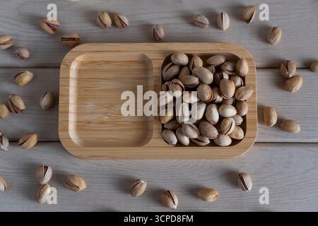 Blick von oben auf rustikale Pistazien auf einem Holztablett, verstreut auf einer hellen Holzoberfläche, die natürliche Texturen und Erdtöne hervorhebt Stockfoto