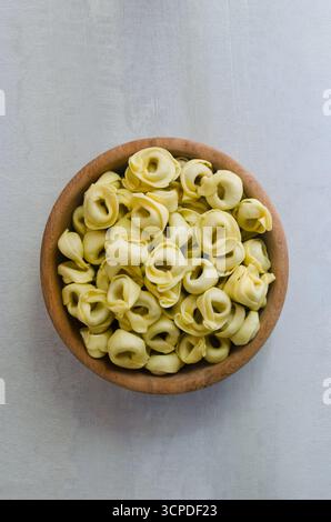 Rohe hausgemachte Tortellini in einer Holzschale auf grauem Hintergrund. Traditionelles italienisches Essenskonzept. Selektiver Fokus. Vertikale Ausrichtung. Draufsicht. Stockfoto