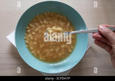 Hausgemachte klare Brühe mit kleinen sternförmigen Nudeln, serviert in einer blauen Schüssel. Warme Suppe mit einem Löffel, traditionelle einfache Mahlzeit, Nahaufnahme auf W Stockfoto