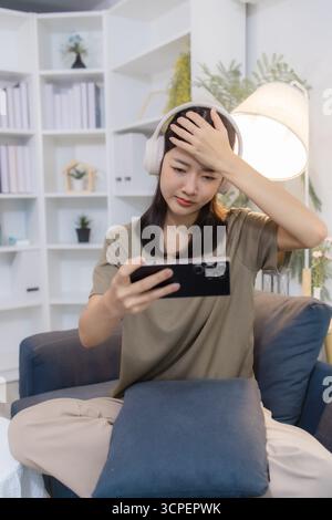Eine junge Frau, die Kopfhörer trägt, sitzt auf einem Sofa, während sie mit ihrem Smartphone interagiert, und zeigt gemischte Emotionen von Freude, Aufregung und Frustration A Stockfoto