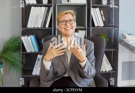 Glückliche aktive Frau, die im Büro sitzt, Gesten gibt, spricht oder erklärt, lächelt und Begeisterung ausdrückt Stockfoto