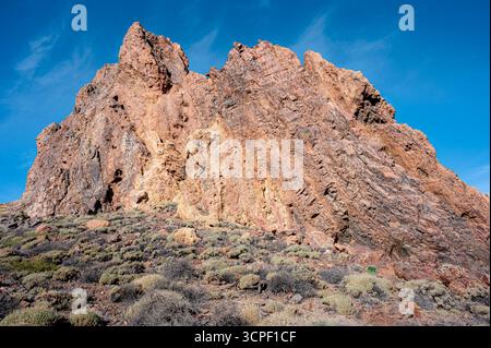 Niedrigwinkelaufnahme eines großen orangen vulkanischen Felsens, der aus Wüstengelände aufsteigt, umgeben von karger Vegetation und einheimischen Pflanzen am Cañadas del Teide Stockfoto