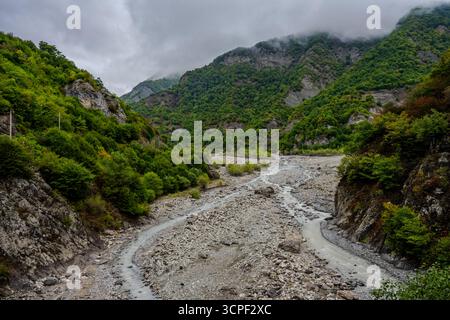 Ein felsiges Flussbett schlängelt sich durch ein steiles Bergtal mit dichten grünen Wäldern unter bewölktem Himmel in Aserbaidschan. Nebel bedeckt die Spitzen in der Distank Stockfoto