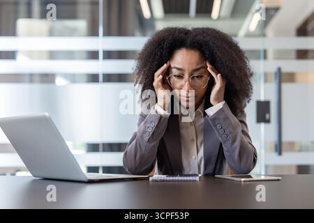 Eine ernste junge afroamerikanische Frau sitzt an ihrem Schreibtisch im Büro und hält nachdenklich ihren Kopf in den Händen. Stockfoto