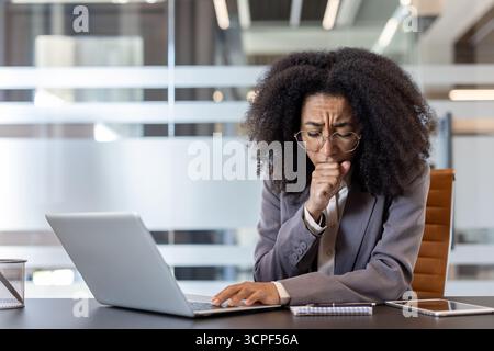 Kranke junge afroamerikanische Geschäftsfrau hustet an ihrem Arbeitsplatz und fühlt sich unwohl. Stockfoto
