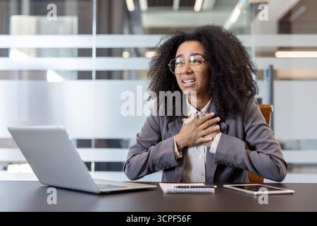 Verängstigte Afroamerikanerin in einem Anzug, die im Büro an ihrem Schreibtisch sitzt, ihre Hände an der Brust hält und eine Panikattacke erlitt. Stockfoto