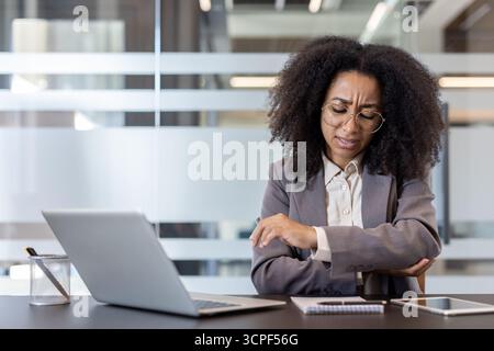 Müde junge afroamerikanische Frau sitzt an einem Schreibtisch im Büro und hält ihren Ellenbogen mit der Hand. Stockfoto