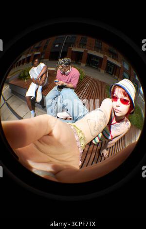 Junge kaukasische Frau mit Tattoos, die Selfie mit Smartphone mit Fischaugenlinse macht, während sie draußen auf der Bank sitzt, Schwarzer und kaukasischer Mann sitzen im Hintergrund Stockfoto