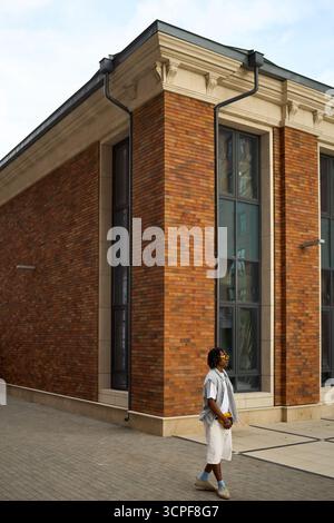 Junger schwarzer Mann, der an der Fassade eines Backsteingebäudes entlang läuft, Sonnenbrille und Tasche trägt, urbane Umgebung mit hohen Fenstern und sichtbaren architektonischen Details Stockfoto