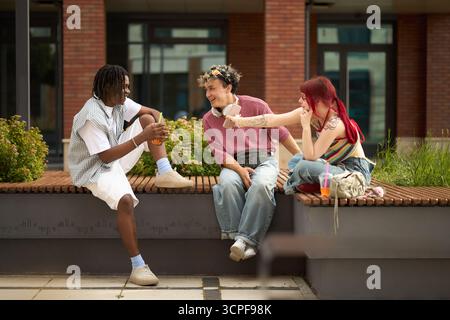 Gruppe von drei Jugendlichen Studenten, Schwarzer Mann und zwei kaukasische Freunde, die draußen auf der Bank sitzen, lachen und reden, während die junge Frau einen Drink hält und zu Freunden gestickt hat Stockfoto
