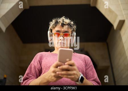 Porträt eines jungen kaukasischen Mannes mit Kopfhörern und Sonnenbrillen im Freien mit Smartphone, mit beiden Händen das Gerät halten und auf den Bildschirm schauen, modernes Technologiekonzept Stockfoto