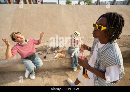 Eine Gruppe multiethnischer Teenager, die sich im Freien entspannen, ein schwarzer Teenager mit Sonnenbrille und Eis essen, während zwei kaukasische Teenager auf einem Betonvorsprung sitzen und Snacks essen und lachen Stockfoto