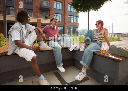 Gruppe von drei Teenagern, schwarz, asiatisch männlich, kaukasisch weiblich, sitzen draußen auf Holzbänken, lachen und unterhalten sich zusammen in der Nähe des modernen Gebäudes und des Yachthafens, genießen Freizeit Stockfoto
