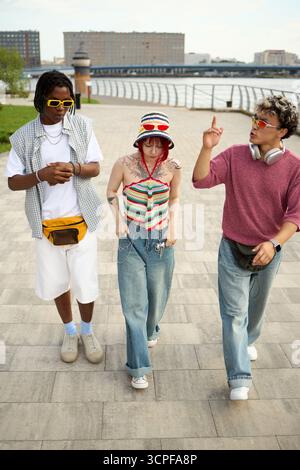 Gruppe von drei jungen Freunden, Schwarzer Mann, Kaukasierin, Kaukasier Mann, spazieren draußen in der Nähe des Ufers, unterhalten sich, tragen legere Kleidung, genießen urbane Umgebung Stockfoto