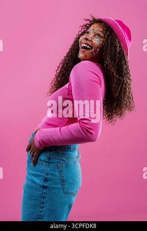 Modeporträt einer fröhlichen jungen Frau mit lockigem Haar, rosa Eimerhut, rosa Langarmshirt und Jeans, lacht vor dem bunten rhein Stockfoto