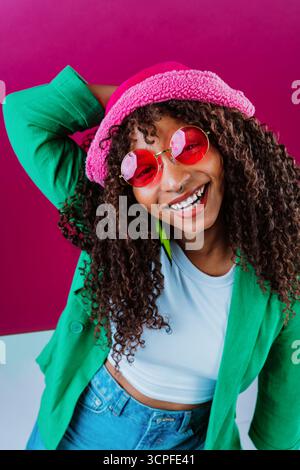 Die modische junge Frau mit lockigen Haaren lächelt hell, während sie einen rosa flauschigen Hut, eine rosa Sonnenbrille und eine grüne Jacke vor einem leuchtenden zwei Tonnen schweren Haar trägt Stockfoto