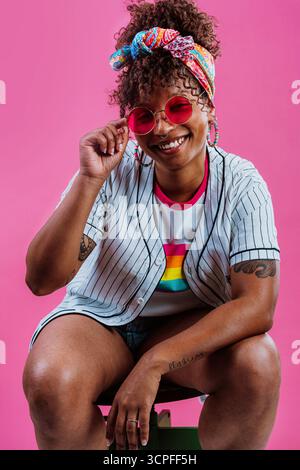 Studio-Porträt einer fröhlichen jungen Frau mit farbenfrohem Kopftuch und Sonnenbrille, die ein Baseballtrikot mit einem Regenbogen-T-Shirt darunter trägt und posiert Stockfoto