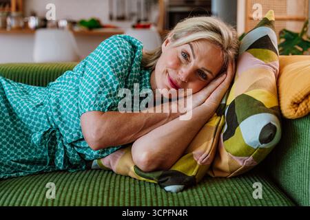 Reife Frau in einem blaugrünen Kleid, die auf einer grünen Couch liegt und ein buntes Kissen umschließt. Das gemütliche Wohnzimmer bietet Entspannung und Komfort in Stockfoto