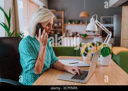 Konzentrierter, reifer freiberuflicher Mitarbeiter, der vom Home Office aus mit Laptop arbeitet und mit dem Mobiltelefon telefoniert, während er an einem Holztisch in einem modernen Apartment sitzt Stockfoto